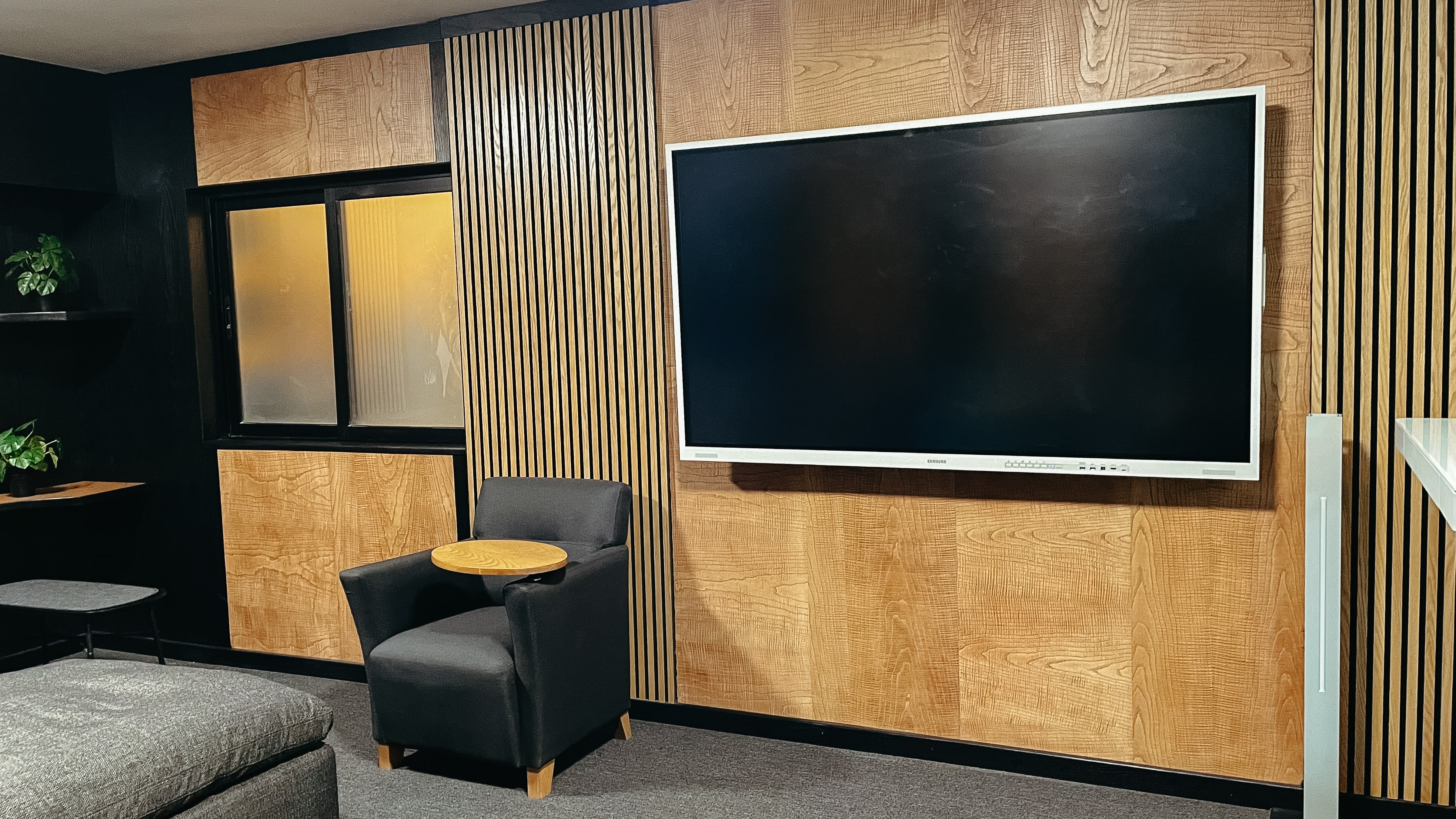 Área de descanso con un sillón gris, una mesa auxiliar de madera y una pantalla grande en la pared de madera.