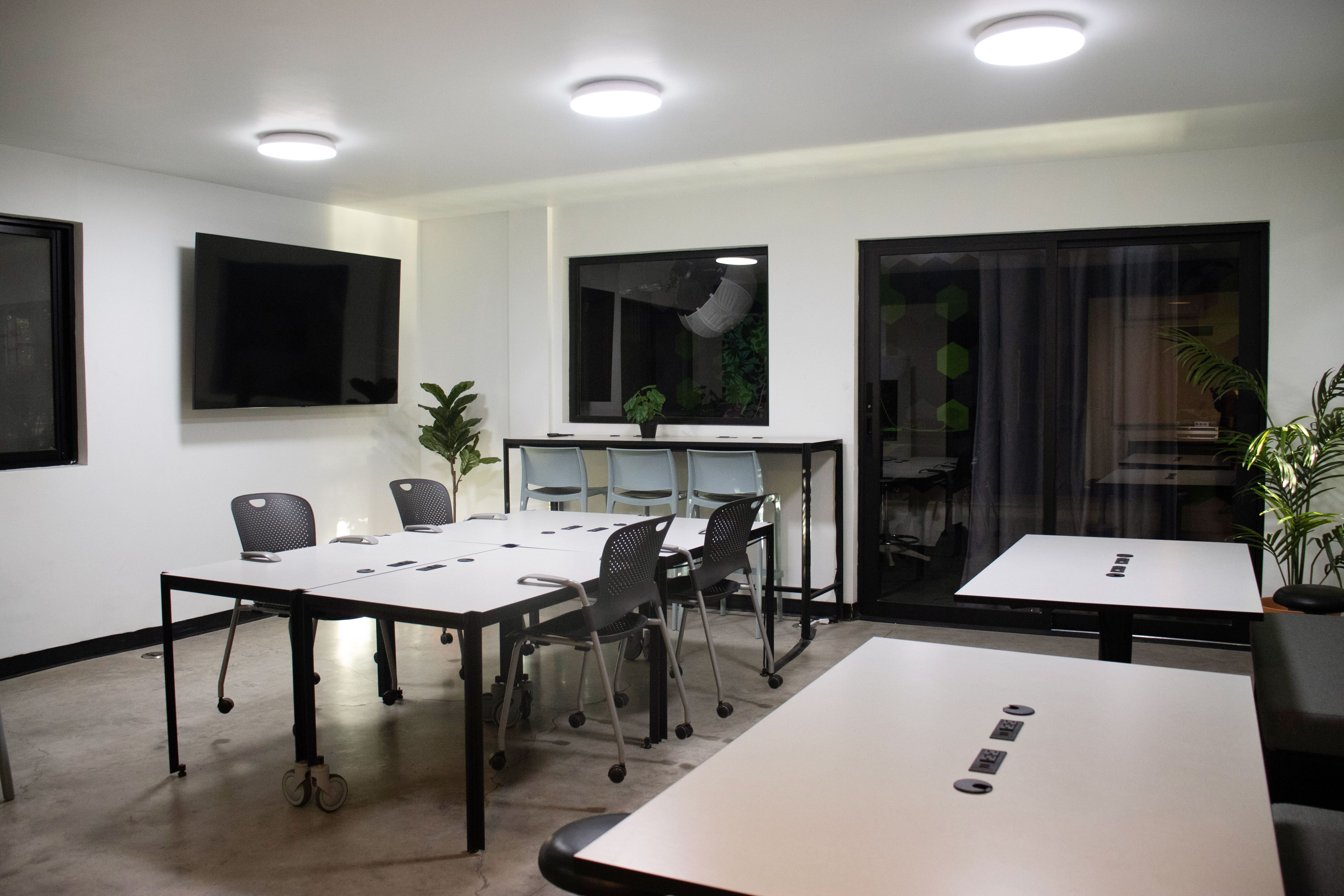Zona de trabajo compartida con mesas blancas, sillas ergonómicas y una pantalla grande, decorada con plantas y un estilo moderno.